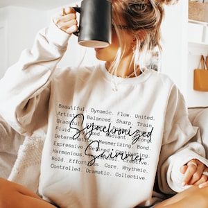 Puede incluir: Una sudadera crema con las palabras "Synchronized Swimmer" impresas en escritura negra. La sudadera también tiene una lista de palabras descriptivas en una fuente más pequeña. Una persona sostiene una taza negra.