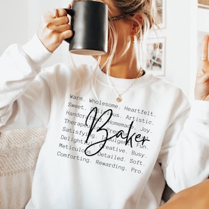 Puede incluir: Sudadera blanca con la palabra "Baker" en escritura negra y una lista de adjetivos que describen el acto de hornear. El texto dice: "Warm. Wholesome. Heartfelt. Sweet. Delicious. Artistic. Handcrafted. Homemade. Joy. Therapeutic. Present. Satisfying. Indulgent. Fun. Delightful. Creative. Busy. Meticulous. Detailed. Soft. Comforting. Rewarding. Pro."