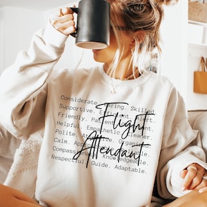 Könnte beinhalten: Ein weißer Pullover mit den Worten "Flight Attendant" in schwarzer Kursivschrift. Die Worte sind von einer Liste positiver Adjektive umgeben, die die Eigenschaften eines Flugbegleiters beschreiben.