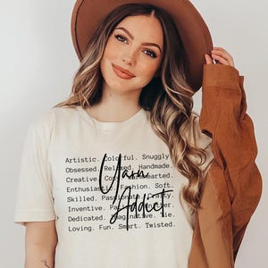 Może przedstawiać: Biały t-shirt z tekstem "Artistic. Colorful. Snuggly. Obsessed. Relaxed. Handmade. Creative. Cozy. Warm-hearted. Enthusiastic. Fashion. Soft. Skilled. Passionate. Crafty. Inventive. Imaginative. Tie. Dedicated.  Loving. Fun. Smart. Twisted." i słowem "Yarn Addict" napisanym kursywą.