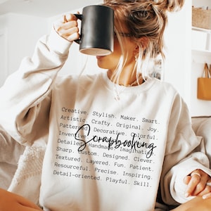 May include: A cream-colored sweatshirt with the word "scrapbooking" in a script font. The sweatshirt has a list of descriptive words in a black font. A person is holding a black mug.