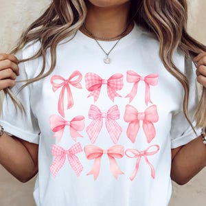 Puede incluir: Camiseta blanca con un dise&ntilde;o de nueve lazos rosas de varios estilos. Los lazos tienen diferentes texturas, incluyendo cuadros y colores lisos. La camiseta es una prenda casual para el d&iacute;a a d&iacute;a.