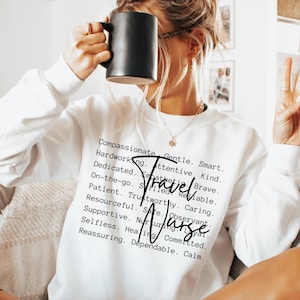 May include: White sweatshirt with the text "Compassionate, Gentle, Smart, Hardworking, Attentive, Kind, Dedicated, Empathetic, Brave, On-the-go, Skilled, Reliable, Travel Nurse, Patient, Trustworthy, Caring, Resourceful, Safe, Observant, Supportive, Nursing, Selfless, Healing, Committed, Reassuring, Dependable, Calm."