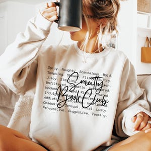 Puede incluir: Una sudadera color crema con el texto "Smutty Book Club" en fuente negra. La sudadera tiene cuello redondo y mangas largas. Una taza negra se sostiene frente a la cara. Las palabras "Spicy, Naughty, Tempting" están impresas encima del texto.