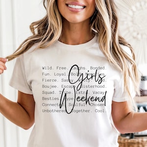 May include: White t-shirt with black text. The text includes the words "Girls Weekend" in a cursive font, surrounded by a list of words like "Wild," "Free," and "Fun." The shirt is worn by a person.