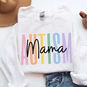 Puede incluir: Sudadera blanca con las palabras "AUTISM Mama" en letras de bloque coloridas. La palabra "Mama" está en una fuente de escritura negra. Un sombrero marrón, una taza de café y unos vaqueros están en el fondo.