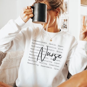 K&ouml;nnte beinhalten: Wei&szlig;er Pullover mit dem Wort "Nurse" in schwarzer Kursivschrift und einer Liste positiver Attribute, die den Beruf beschreiben. Der Text lautet: Mitf&uuml;hlend, Sanft, Klug, Flei&szlig;ig, Aufmerksam, Freundlich, Engagiert, Empathisch, Mutig, Belastbar, Geschickt, Zuverl&auml;ssig, Geduldig, Vertrauensvoll, Beobachtungsgabe, Einfallsreich, Held, Loyal, Unterst&uuml;tzend, F&uuml;rsorglich, Selbstlos, Heilend, Engagiert, Beruhigend, Zuverl&auml;ssig, Ruhig.