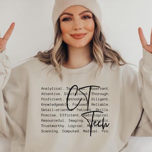 Puede incluir: Una sudadera beige con la palabra "Tech" en una fuente estilizada, acompañada de una lista de palabras descriptivas. La persona de la imagen lleva un gorro beige y hace un signo de la paz con los dedos.