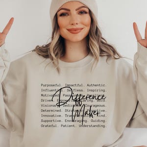 May include: A beige sweatshirt with the words "Difference Maker" and a list of positive attributes printed in black. The person in the image is wearing a beige beanie and making a peace sign with their fingers.