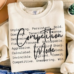 May include: A beige sweatshirt with the text "Competitive Mode" printed in black. The text is surrounded by a list of words describing competitive traits, such as "unyielding", "persistent", "bold", "dominant", "tenacious", "intense", "determined", "focused", "unstoppable", "aggressive", "strategic", "sharp", "calculated", "hungry", "relentless", "invincible", "vigilant", "commit", "competitive", and "unwavering".