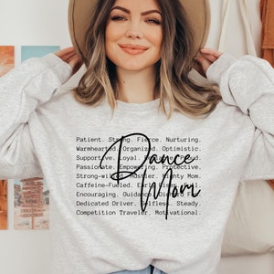 May include: A grey sweatshirt with the text "Patient. Strong. Fierce. Nurturing. Warmhearted. Organized. Optimistic. Supportive. Loyal. Dance Mom. Passionate. Empowering. Protective. Strong-willed. Hustler. Mighty Mom. Caffeine-Fueled. Early Riser. Encouraging. Guidance. Dedicated Driver. Selfless. Steady. Competition Traveller. Motivational." printed on the front.