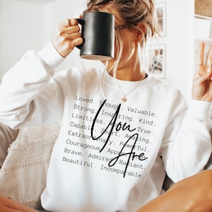 May include: White sweatshirt with the text "You Are" in black cursive lettering. The text is surrounded by a list of positive adjectives, including "Loved", "Enough", "Valuable", "Strong", "Inspiring", "Kind", "Limitless", "True", "Capable", "Smart", "Amazing", "Extraordinary", "Powerful", "Courageous", "Appreciated", "Brave", "Admired", "Radiant", and "Beautiful".