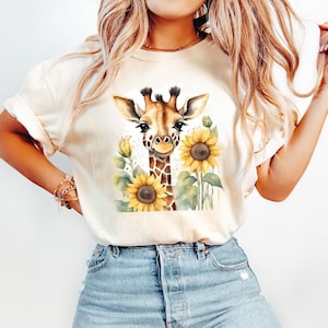 May include: A white t-shirt featuring a watercolour illustration of a giraffe with sunflowers. The giraffe is brown and has a long neck and spots. The sunflowers are yellow with brown centres.