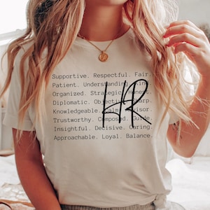 May include: White t-shirt with black text that reads "Supportive. Respectful. Fair. Patient. Understanding. Kind. Organized. Strategic. Honest. Diplomatic. Objective. Sharp. HR. Knowledgeable. Calm. Advisor. Trustworthy. Composed. Guide. Insightful. Decisive. Caring. Approachable. Loyal. Balance."