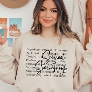 Puede incluir: Una sudadera beige con las palabras "School Secretary" en una fuente manuscrita. La sudadera también tiene una lista de atributos positivos como "Organizado", "Paciente" y "Amable" impresos en negro.