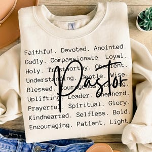 May include: A cream-coloured sweatshirt featuring the word "Pastor" in a script font. The shirt is printed with a list of positive attributes including "Faithful," "Devoted," and "Kindhearted." The sweatshirt is paired with blue jeans and a tan hat.