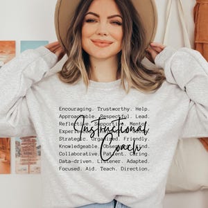May include: A light gray sweatshirt with the words "Instructional Coach" printed in black. The sweatshirt also has a list of positive attributes. The model is wearing a tan hat and blue jeans.