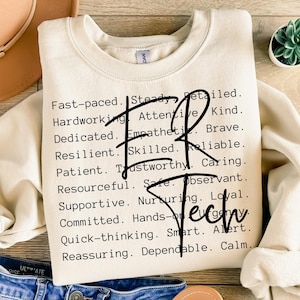 Könnte beinhalten: Ein beiger Pullover mit einem schwarzen grafischen Druck einer Liste positiver Persönlichkeitsmerkmale. Der Text enthält Wörter wie "schnelllebig", "beständig", "detailliert", "fleißig", "aufmerksam", "freundlich", "engagiert", "empathisch", "mutig", "widerstandsfähig", "geschickt", "zuverlässig", "geduldig", "vertrauenswürdig", "fürsorglich", "erfinderisch", "sicher", "beobachtend", "unterstützend", "fürsorglich", "loyal", "engagiert", "praktisch", "schnell denkend", "klug", "wachsam", "beruhigend", "zuverlässig" und "ruhig".