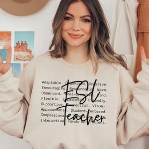 May include: Beige sweatshirt with the word "Teacher" in a script font, with the letters "FSJ" above it. The sweatshirt has a list of positive adjectives. The person is smiling and giving a peace sign.