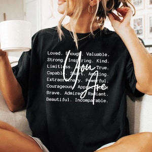 May include: Black t-shirt with white text that reads "You Are" and a list of positive attributes, including loved, enough, valuable, strong, inspiring, kind, limitless, true, capable, smart, amazing, extraordinary, powerful, courageous, appreciated, brave, admired, radiant, and beautiful.