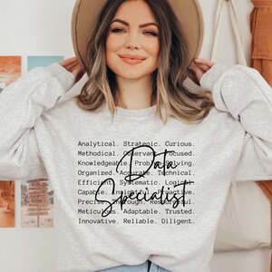 Könnte beinhalten: Ein hellgraues Sweatshirt mit dem Schriftzug "Data Specialist" in einer stilvollen Schriftart. Der Text wird von einer Liste beschreibender Wörter wie "Analytical", "Strategic" und "Curious" begleitet. Das Sweatshirt wird von einer Person getragen.
