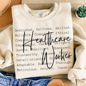 May include: A beige sweatshirt with the words "Healthcare Worker" printed in black. The words are surrounded by a list of positive adjectives describing the qualities of a healthcare worker.