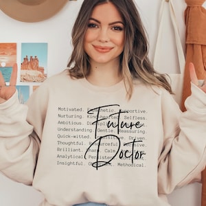 May include: A light beige sweatshirt with the text "Future Doctor" in black lettering. The text is surrounded by a list of positive adjectives describing a person's character.