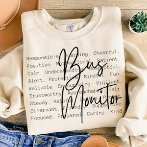 May include: Cream-colored sweatshirt with the words "Bus Monitor" in a black script font. The sweatshirt also features a list of positive attributes. The sweatshirt is paired with blue jeans and a tan hat.