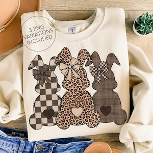 Puede incluir: Sudadera crema con tres siluetas de conejos con diseños estampados: cuadros, estampado de leopardo y cuadros escoceses. Cada conejo tiene un lazo y un corazón. El texto "3 PNG VARIATIONS INCLUDED" está en un círculo.