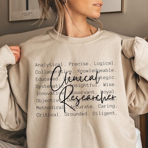 May include: A beige sweatshirt with the text "Clinical Researcher" in black lettering. The text is surrounded by a list of positive adjectives describing the qualities of a clinical researcher.