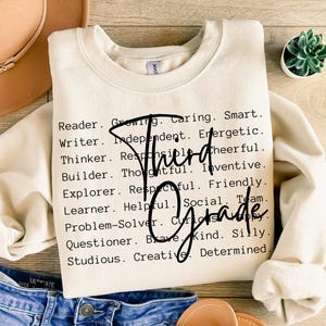 Może przedstawiać: Kremowa bluza z napisem "Third Grade" czcionką w kolorze czarnym. Bluza ma listę pozytywnych przymiotnik&oacute;w, takich jak "Reader", "Caring" i "Creative" wydrukowanych na czarno.