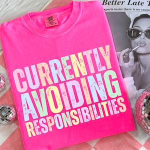 May include: A bright pink crew neck sweatshirt with the phrase "CURRENTLY AVOIDING RESPONSIBILITIES" printed in pastel colors. The sweatshirt is on a pink and white checkered surface, with a disco ball and a newspaper in the background.