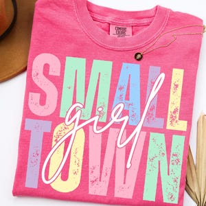 May include: A pink Comfort Colors t-shirt with the words "SMALL TOWN girl" in a distressed, multi-colored font. A gold necklace with a black pendant rests on the shirt. A brown hat is partially visible in the upper left corner.