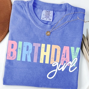 Może przedstawiać: Niebieska koszulka Comfort Colors z napisem "BIRTHDAY girl" w kolorowych literach blokowych. Złoty naszyjnik z małą zawieszką spoczywa na koszulce. Koszulka w rozmiarze S-P-CH.