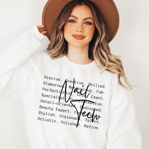 May include: White sweatshirt with black text that reads "Precise. Creative. Skilled. Glamorous. Meticulous. Fab. Perfectionist. Chic. Exact. Specialist. Transformative. Detail-oriented. Dedicated. Beauty Expert. Fashionable. Stylish. Visionary. Talent. Artistic. Polished. Refine."