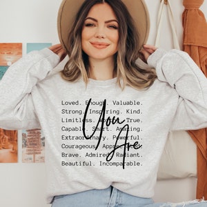 May include: A gray sweatshirt with the text "Loved. Enough. Valuable. Strong. Inspiring. Kind. Limitless. You Are. Capable. Smart. Amazing. Extraordinary. Powerful. Courageous. Appreciated. Brave. Admired. Radiant. Beautiful. Incomparable."