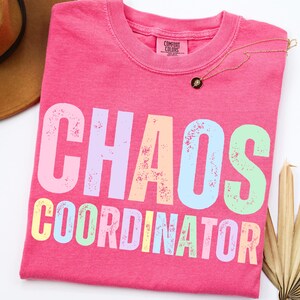 Peut inclure: Un t-shirt rose vif avec les mots "CHAOS COORDINATOR" imprimés en couleurs pastel. Le t-shirt a un col rond et une étiquette Comfort Colors. Un collier en or avec un pendentif noir est drapé sur le t-shirt.
