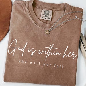 May include: A brown Comfort Colors t-shirt with the text "God is within her, she will not fall" in white script. A silver necklace with a small pendant rests on the shirt. The shirt is folded and displayed on a white surface.
