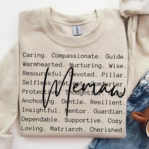 May include: A beige sweatshirt with the word "Woman" in a cursive font. The sweatshirt is printed with a list of words describing a woman, including "Caring," "Compassionate," and "Loving."