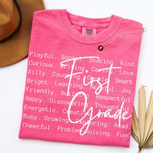 May include: A pink Comfort Colors t-shirt with the words "First Grade" in white script. The shirt is adorned with a gold necklace and a list of positive adjectives. A brown hat is partially visible in the background.