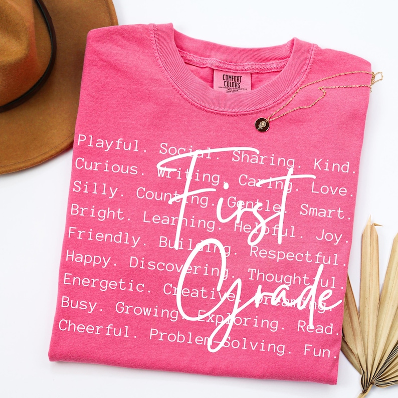 May include: A pink Comfort Colors t-shirt with the words "First Grade" in white script. The shirt is adorned with a gold necklace and a list of positive adjectives. A brown hat is partially visible in the background.