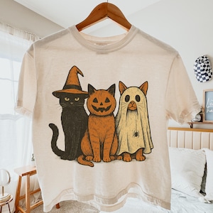 K&ouml;nnte beinhalten: Cremefarbenes T-Shirt mit einem Halloween-Motiv von drei Katzen. Eine Katze tr&auml;gt einen Hexenhut, eine andere ist ein K&uuml;rbis und die dritte ist ein Geist. Das Design verwendet Schwarz-, Orange- und Cremefarben.