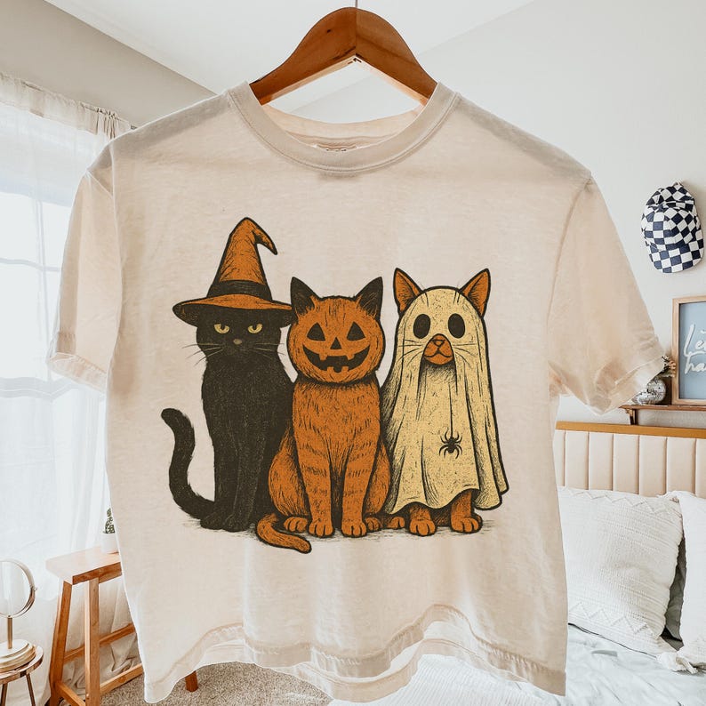K&ouml;nnte beinhalten: Cremefarbenes T-Shirt mit einem Halloween-Motiv von drei Katzen. Eine Katze tr&auml;gt einen Hexenhut, eine andere ist ein K&uuml;rbis und die dritte ist ein Geist. Das Design verwendet Schwarz-, Orange- und Cremefarben.