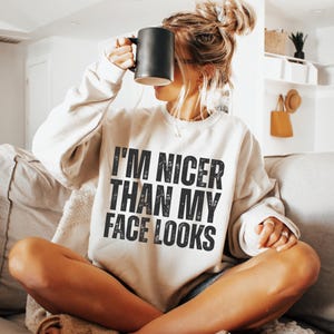 Pu&ograve; includere: Felpa color crema con la scritta "I'M NICER THAN MY FACE LOOKS" in nero. La persona tiene in mano una tazza nera. La felpa &egrave; uno stile casual e confortevole.