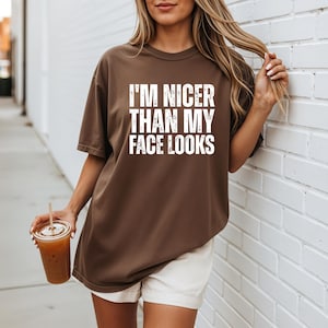 Pu&ograve; includere: Una maglietta marrone con la scritta "I'M NICER THAN MY FACE LOOKS" a blocchi bianchi. La maglietta &egrave; indossata con pantaloncini bianchi. Una persona tiene in mano un bicchiere di plastica trasparente con una cannuccia e una bevanda marrone.