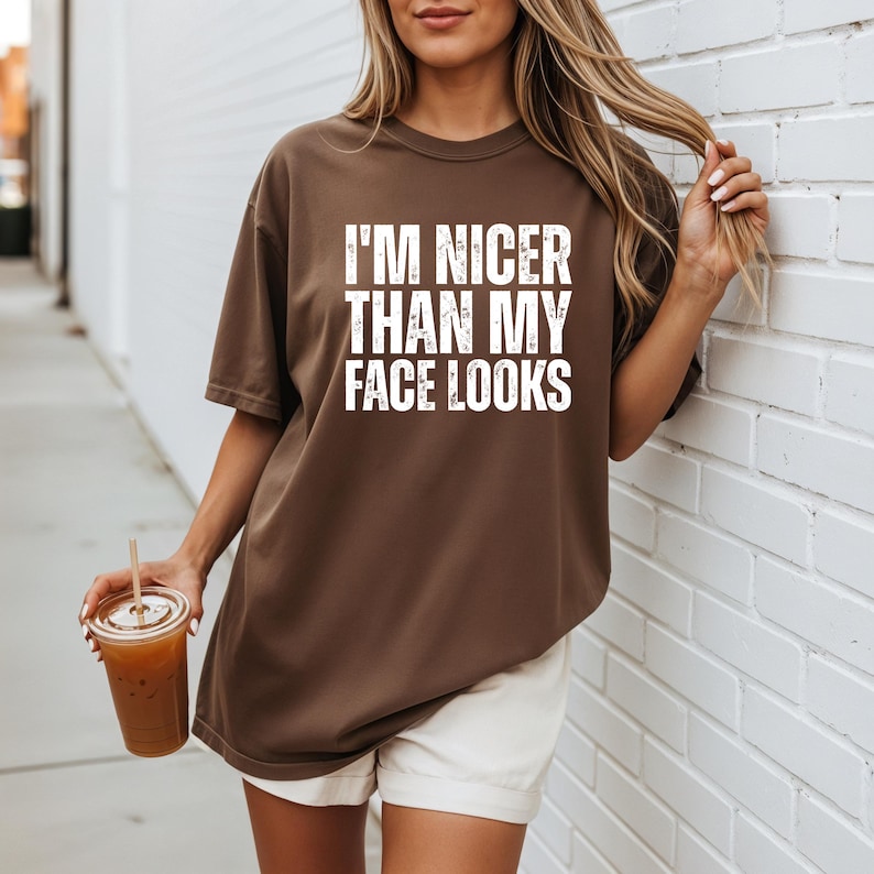 Pu&ograve; includere: Una maglietta marrone con la scritta "I'M NICER THAN MY FACE LOOKS" a blocchi bianchi. La maglietta &egrave; indossata con pantaloncini bianchi. Una persona tiene in mano un bicchiere di plastica trasparente con una cannuccia e una bevanda marrone.
