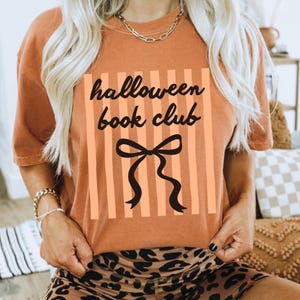 May include: An orange t-shirt with the text "halloween book club" in black script, set against a background of vertical orange stripes. A black bow graphic is centered below the text. The shirt is a casual style.