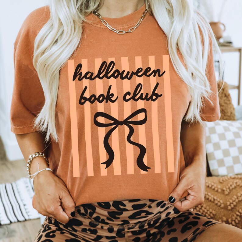 May include: An orange t-shirt with the text "halloween book club" in black script, set against a background of vertical orange stripes. A black bow graphic is centered below the text. The shirt is a casual style.