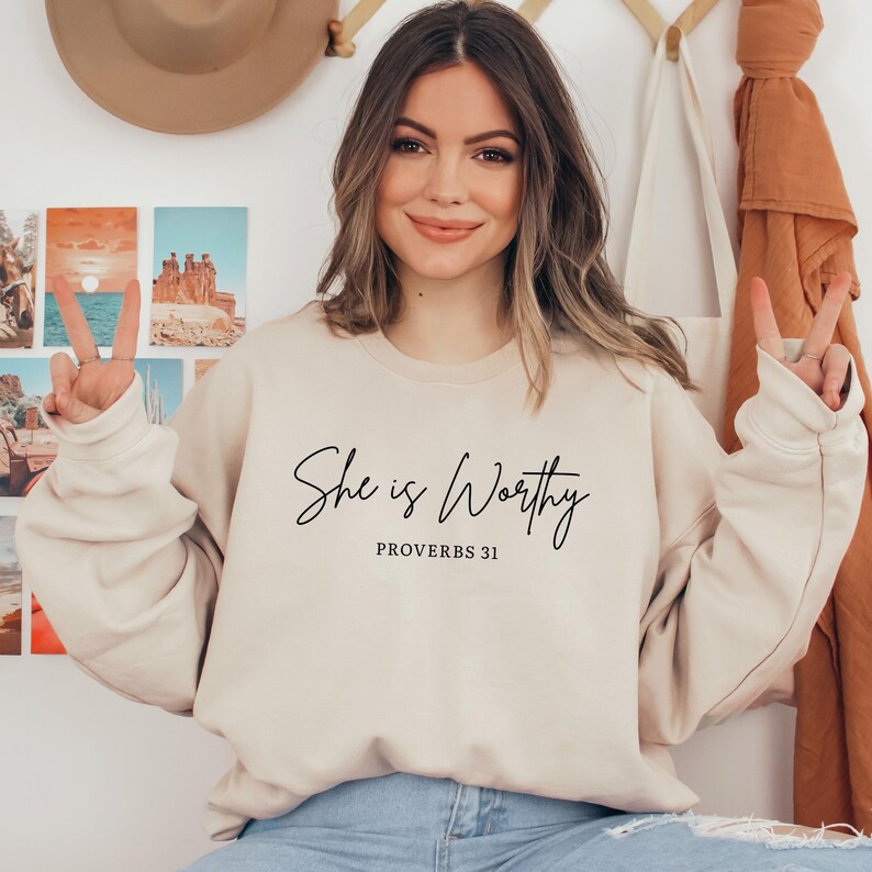 May include: A beige sweatshirt with the text "She is Worthy" in a black script font. Below the text is "Proverbs 31". The sweatshirt is worn by a person.