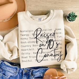 Puede incluir: Sudadera de color crema con la frase "Raised on 90s Country" en fuente negra. La sudadera tiene cuello redondo y mangas largas. Palabras adicionales relacionadas con la música country están impresas alrededor del texto principal.